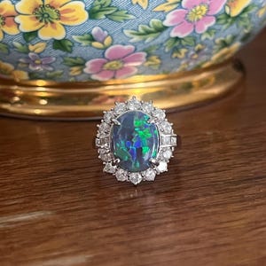 Puede incluir: Un anillo de plata con una gran opala negra ovalada rodeada de diamantes más pequeños. La opala tiene un brillo iridiscente verde y azul.