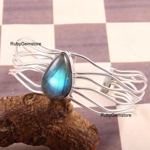 Könnte beinhalten: Silbernes Armreif-Armband mit einem tropfenförmigen Labradorit-Edelstein. Der Edelstein hat einen blauen und grünen irisierenden Schimmer.