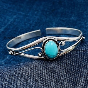 Könnte beinhalten: Silbernes Armreif-Armband mit einem Türkis-Stein, der in einem dekorativen silbernen Rahmen gefasst ist. Das Armband liegt auf einem blauen Jeans-Hintergrund.