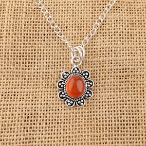 May include: A silver-toned necklace featuring a pendant with a central oval-shaped orange stone. The stone is surrounded by a decorative metal frame with a scalloped edge and small circular bead accents, suspended from a simple silver-toned chain.