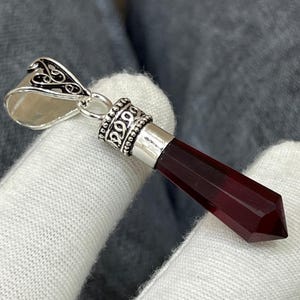 Könnte beinhalten: Ein silberner Anhänger mit einem facettierten, dunkelroten Kristall. Der Anhänger hat eine dekorative silberne Öse und eine silberne Kappe an der Oberseite des Kristalls. Der Kristall hat eine tiefe, transluzente rote Farbe.