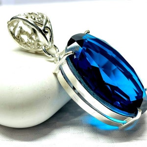 Könnte beinhalten: Anhänger mit einem großen, rechteckigen, tiefblauen Edelstein in Silber gefasst. Der Edelstein ist facettiert und fängt das Licht ein. Die silberne Öse hat ein aufwendiges, durchbrochenes Design. Das Design des Anhängers ist elegant.