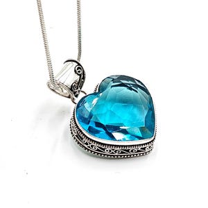 Könnte beinhalten: Halskette mit herzförmigem Anhänger und einem großen, facettierten, hellblauen Edelstein. Der Edelstein ist in einen silberfarbenen Metallrahmen mit einem verzierten Filigran-Design gefasst. Die Halskette hat eine dünne, silberfarbene Schlangenkette.