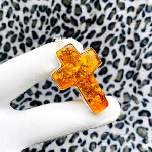 Könnte beinhalten: Ein silberner Anhänger mit einem Kreuz aus Bernstein. Der Bernstein hat einen warmen, gold-orangenen Farbton mit sichtbaren Einschlüssen. Das Kreuz ist in einen silbernen Rahmen gefasst und hat eine Schlaufe für eine Kette.