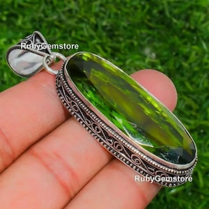 Könnte beinhalten: Ein silberner Anhänger mit einem ovalen, grünen Edelstein, der in einen silbernen Filigranrahmen gefasst ist. Der Anhänger befindet sich auf einer Hand.