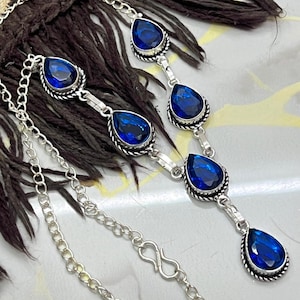 Könnte beinhalten: Eine silberne Halskette mit tropfenförmigen, tiefblauen Edelsteinen. Die Edelsteine sind in einem dekorativen Silberrahmen gefasst und durch filigrane Silberglieder verbunden. Die Halskette hat ein klassisches Design.