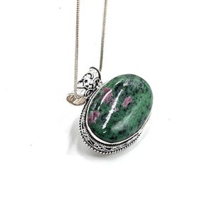 Könnte beinhalten: Silberkette mit einem ovalen Anhänger. Der Anhänger hat einen grünen Stein mit roten Einschlüssen, gefasst in einem detaillierten Silberrahmen. Die Kette ist ein filigranes Schlangenkettendesign.