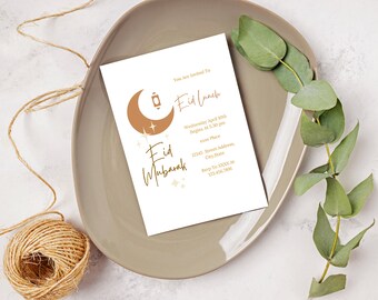 May include: A white invitation card with gold text and graphics for an Eid Lunch. The card features a crescent moon, lantern, and stars. The card is on a gray plate with a ball of twine and eucalyptus leaves.