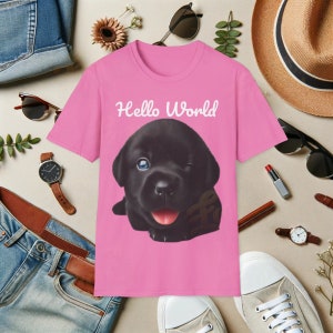May include: Pink t-shirt featuring a black puppy wearing a brown collar. The puppy is winking and has its tongue sticking out. The text "Hello World" is printed in white on the shirt.