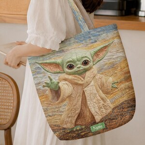 Puede incluir: Un bolso de tela rectangular con una ilustraci&oacute;n impresa de una peque&ntilde;a criatura verde de orejas grandes que viste una t&uacute;nica color canela. El fondo tiene un patr&oacute;n texturizado de pinceladas en tonos azul, amarillo y marr&oacute;n. Una peque&ntilde;a etiqueta rectangular verde con la palabra Name se encuentra en la parte inferior.