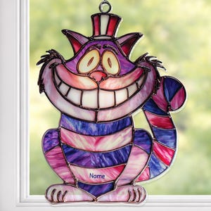 May include: Stained glass Cheshire Cat suncatcher with a wide grin, striped body, and top hat. The cat is purple, pink, and blue, with a chain for hanging. The word "Name" is printed on the cat's body.