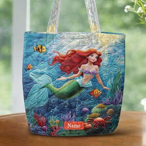 Puede incluir: Un bolso tote acolchado con una vibrante escena submarina que muestra a una sirena de largo cabello rojo, cola verde y top de conchas moradas. El dise&ntilde;o incluye peces tropicales coloridos, corales y una etiqueta rectangular roja con la palabra Name.
