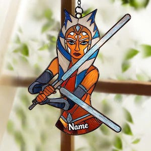 May include: A stained glass ornament featuring a character with orange skin, blue and white markings, and blue lightsabers. The character is holding two lightsabers. The ornament has a chain for hanging and the word "Name" is visible.