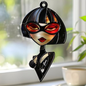 May include: A black and red decorative hanging ornament featuring a stylized female character with a bob haircut and red-tinted glasses. The ornament is suspended by a silver chain. The word "Alane" is written on the ornament.