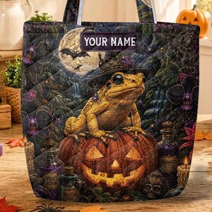 May include: A quilted tote bag featuring a graphic print of a yellow frog wearing a witch hat while sitting on a jack-o'-lantern. The design includes a full moon, spiderwebs, and potion bottles. A rectangular label at the top reads YOUR NAME.