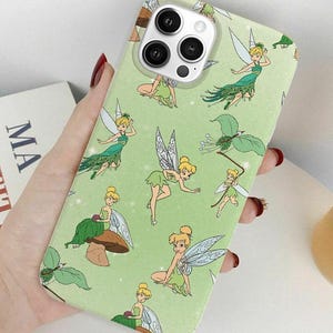 May include: A light green phone case featuring a repeating pattern of cartoon fairies in various poses. The fairies are depicted with blonde hair, green dresses, and wings. The case is held in a person's hand, with a book and a cup visible in the background.