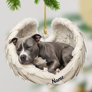 May include: A flat, circular hanging ornament featuring a gray and white pit bull puppy sleeping inside a pair of large, white feathered angel wings. The ornament includes a gold hanging string and the black text Name printed on the lower right.