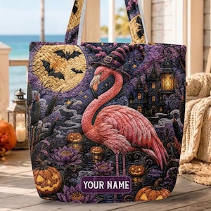 May include: A quilted tote bag featuring a pink flamingo wearing a witch hat, set against a purple night scene with a full moon, bats, jack-o'-lanterns, and a dark house. The bag includes a rectangular label with the text YOUR NAME.