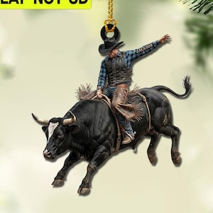 Op de afbeelding: Een plat 2D-hangornament met een stierenrijder in een zwarte hoed, geruit overhemd en leren chaps op een zwarte stier met een witte vlek op het voorhoofd. De tekst 2D FLAT-NOT 3D is linksboven zichtbaar. Het ornament hangt aan een gouden koord.