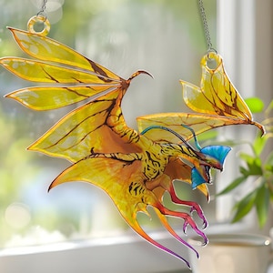 May include: A stained glass dragon suncatcher with yellow, orange, and blue accents. The dragon has outstretched wings and is suspended by a chain. The dragon's wings are a translucent yellow with brown veins.