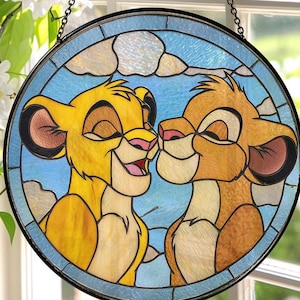 May include: Round stained glass window featuring two lion cubs from The Lion King. The cubs are depicted with closed eyes and smiling, set against a blue sky with white clouds. The window is framed in black and hangs from a chain.