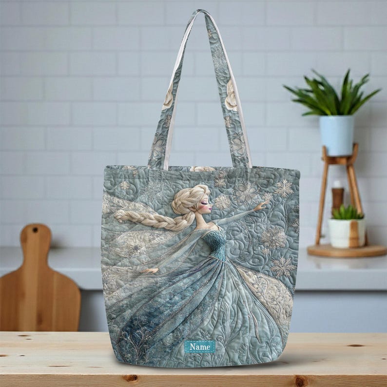 May include: A quilted tote bag in shades of blue and white featuring a printed illustration of a character with long blonde hair in a flowing gown. The design includes snowflake patterns and a small rectangular label with the text Name.