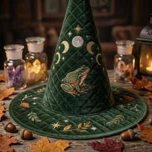 May include: A dark green quilted velvet hat featuring an embroidered frog at the center, surrounded by gold-stitched moon phases, stars, and leaf motifs. The hat has a pointed crown and a wide brim, with detailed stitching throughout the fabric.