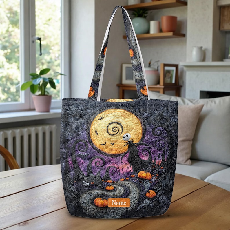 May include: A quilted tote bag featuring a dark, swirling night sky with a large yellow moon, a skeletal figure, and orange pumpkins. The design includes a path leading through a landscape with trees and a rectangular patch labeled Name.