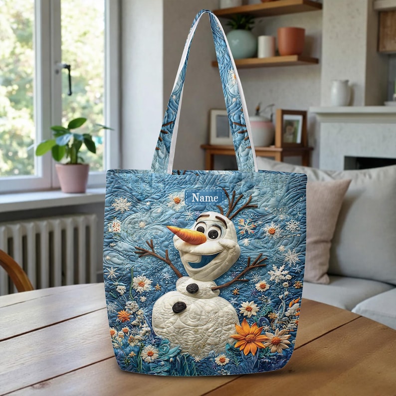 May include: A blue quilted tote bag featuring a central graphic of a snowman with an orange carrot nose, twig arms, and black buttons. The background displays a winter scene with white snowflakes and orange flowers. A rectangular label reads Name.
