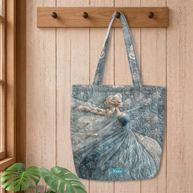 May include: A quilted tote bag featuring a light blue and white illustration of a character with long blonde hair in a flowing gown, surrounded by snowflake patterns. The bag includes a rectangular label with the printed text Name.