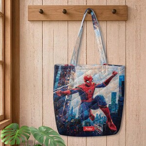May include: A quilted tote bag featuring a graphic print of the superhero Spider-Man in a dynamic pose against a city skyline. The bag has two shoulder straps and a small rectangular red label at the bottom with the word Name printed in white.