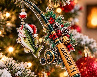Puede incluir: Un adorno navideño plano 2D con forma de caña de pescar con una lubina que lleva un gorro de Papá Noel. La caña presenta ramas de pino, piñas, un lazo de cuadros rojos, una campana dorada y el texto JACKSON 2026 con un icono de pez.