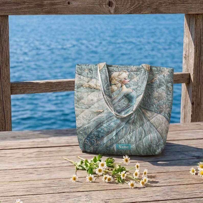May include: A quilted tote bag featuring a light blue and white illustration of a blonde character in a gown with snowflake patterns. The bag has two fabric handles and a small rectangular label at the bottom center with the word Name printed.