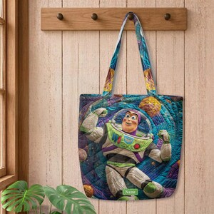 以下が含まれることがあります： 白、緑、紫の宇宙服を着たバズ・ライトイヤーが描かれた、カラフルな宇宙柄のキルティングトートバッグ。背景には惑星が浮かぶ青と紫の渦巻く宇宙模様がデザインされている。緑色の小さなラベルにはNameと記されている。