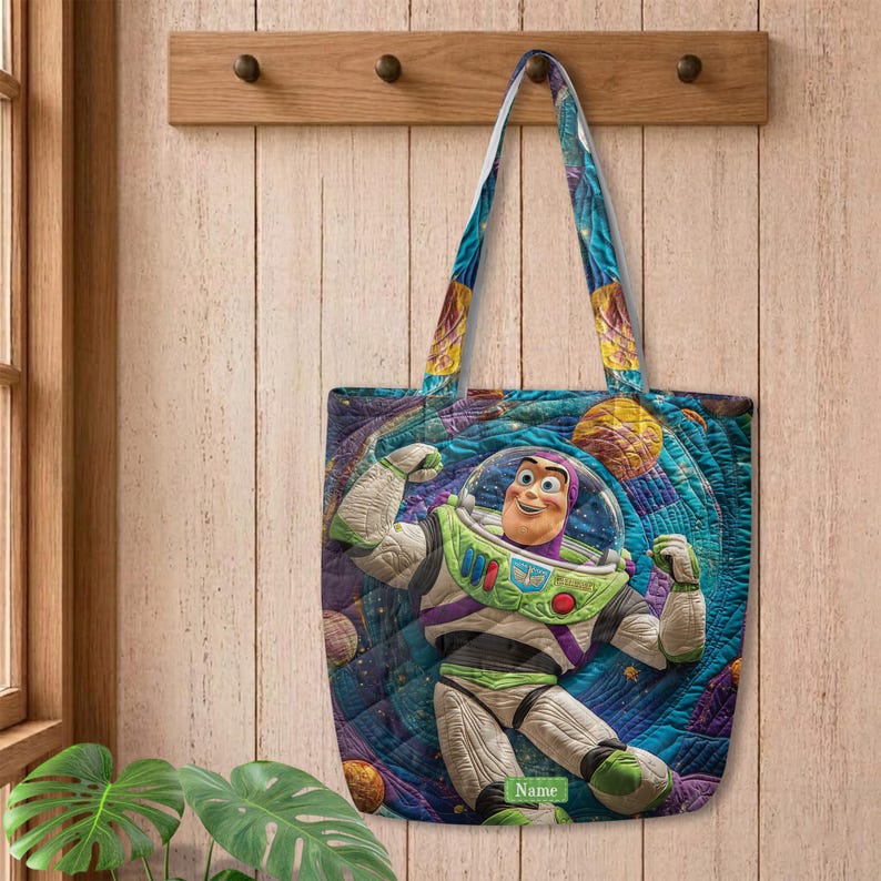 以下が含まれることがあります： 白、緑、紫の宇宙服を着たバズ・ライトイヤーが描かれた、カラフルな宇宙柄のキルティングトートバッグ。背景には惑星が浮かぶ青と紫の渦巻く宇宙模様がデザインされている。緑色の小さなラベルにはNameと記されている。