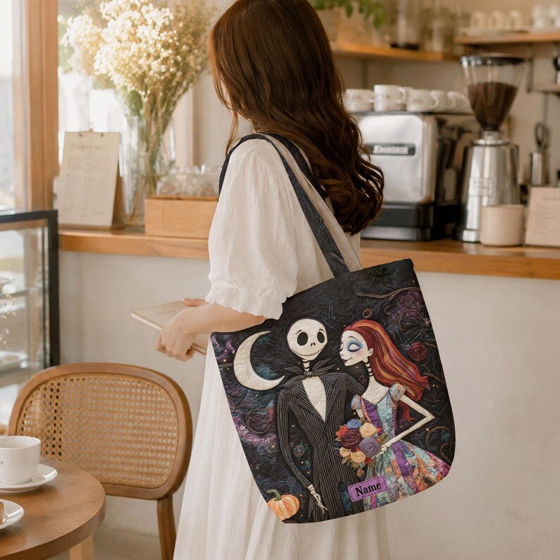 May include: A black tote bag featuring a printed illustration of a skeletal character in a pinstripe suit and a female character with long red hair in a patchwork dress, holding a bouquet of flowers, with a crescent moon and a pumpkin below.