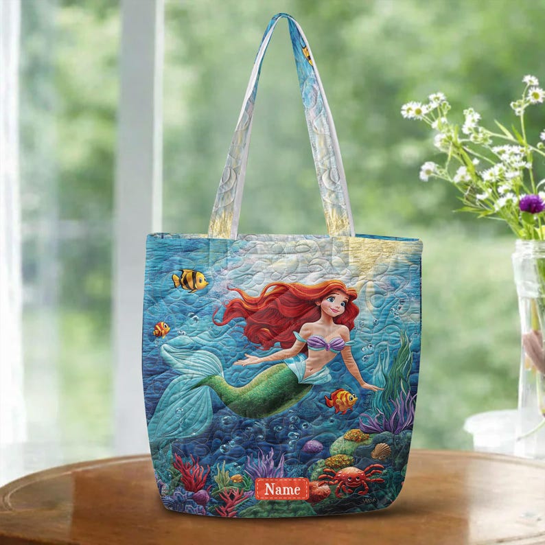 Puede incluir: Un bolso tote acolchado con una vibrante escena submarina que muestra a una sirena de largo cabello rojo, cola verde y top de conchas moradas. El dise&ntilde;o incluye peces tropicales coloridos, corales y una etiqueta rectangular roja con la palabra Name.