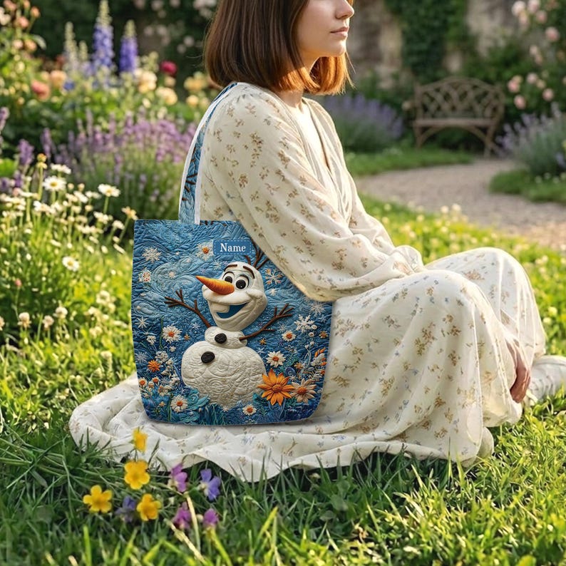 May include: A blue quilted tote bag featuring a central appliqu&eacute; of a snowman with a carrot nose and stick arms, surrounded by embroidered flowers and swirling patterns. A small rectangular label at the top displays the word Name.