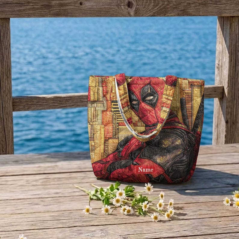 K&ouml;nnte beinhalten: Eine gesteppte Tragetasche mit einem Grafikdruck der Figur Deadpool in Rot und Schwarz vor einem gelben und orangefarbenen geometrischen Hintergrund. Die Tasche enth&auml;lt unten in der Mitte einen rechteckigen roten Aufn&auml;her mit dem Wort Name in Wei&szlig;.
