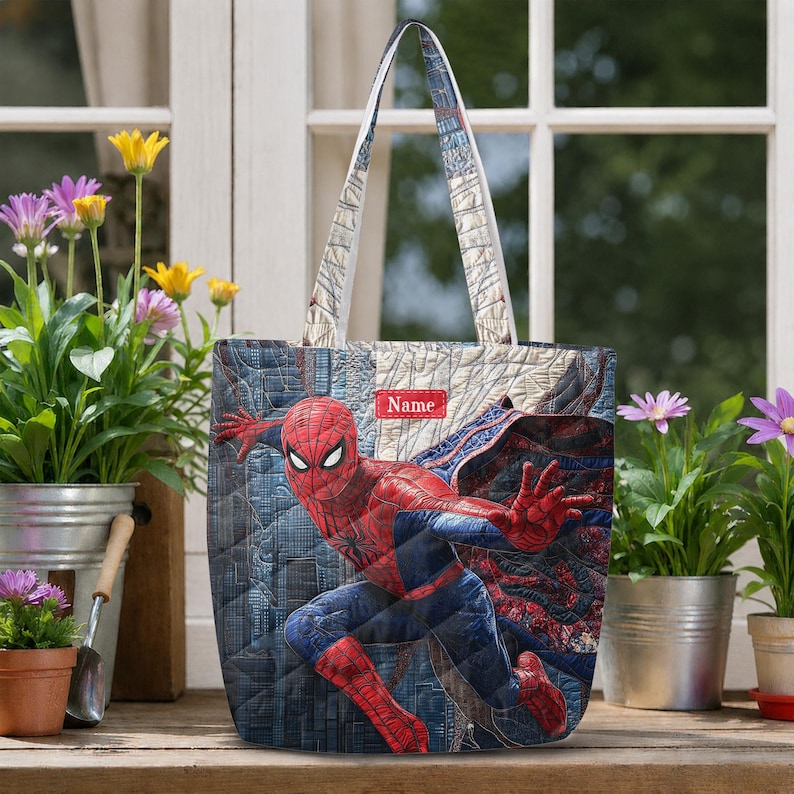 May include: A quilted tote bag featuring a graphic of the character Spider-Man in a dynamic pose. The bag has a blue and red color scheme with a city skyline pattern and a rectangular red patch at the top center with the white text Name.
