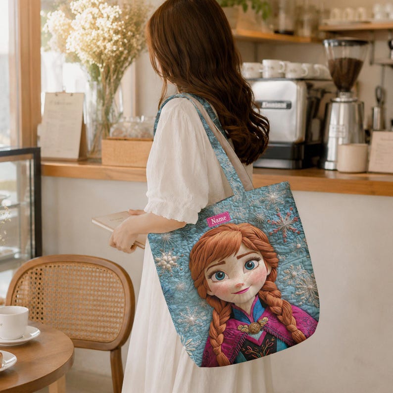 May include: A tote bag featuring a printed graphic of a cartoon character with auburn hair in braids, wearing a magenta and blue outfit. The background of the print is light blue with white snowflake patterns and a small pink rectangle labeled Name.