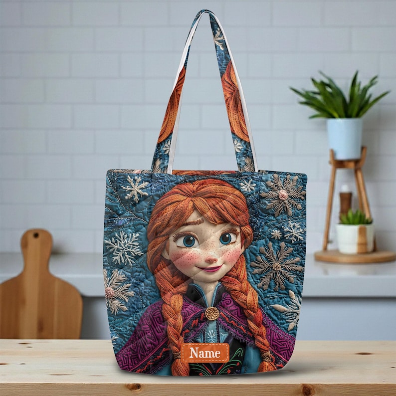 May include: A quilted tote bag featuring a printed illustration of a character with red braided hair, wearing a purple and blue outfit. The background is a dark blue quilted pattern with white snowflake designs. A rectangular tag reads Name.