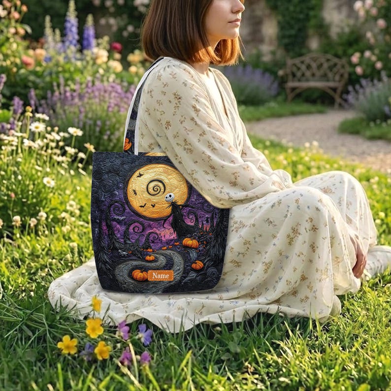 May include: A tote bag featuring a dark, stylized illustration of a winding path, orange pumpkins, a large yellow moon, and a skeletal figure. The design includes a rectangular orange label with the text Name printed in black letters.