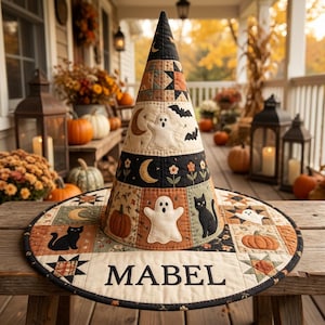 May include: A conical fabric witch hat featuring a patchwork design with Halloween motifs including ghosts, black cats, pumpkins, stars, and moons. The wide brim is embroidered with the name MABEL in black capital letters on a cream background.