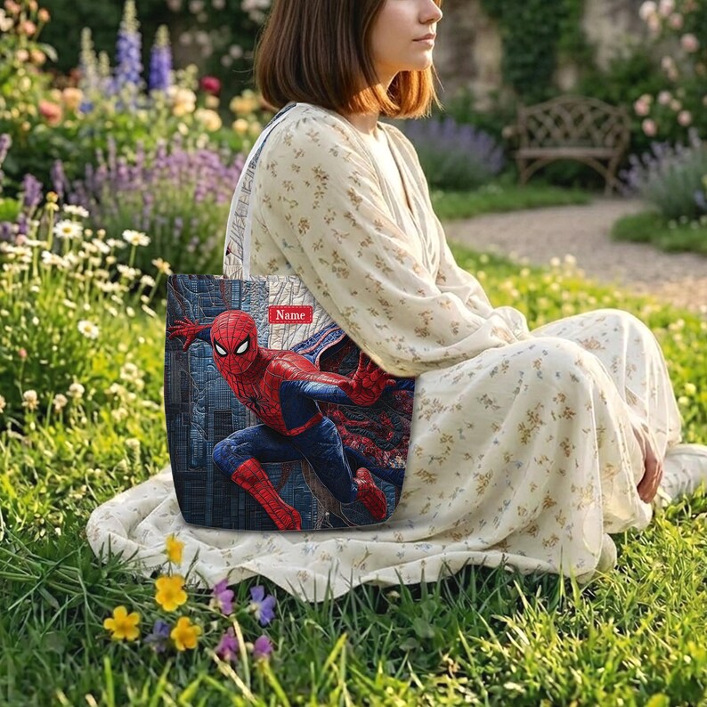 May include: A rectangular tote bag featuring a graphic print of the superhero Spider-Man in a dynamic pose against a dark, textured background. The bag includes a small red rectangular label at the top with the word Name printed in white.
