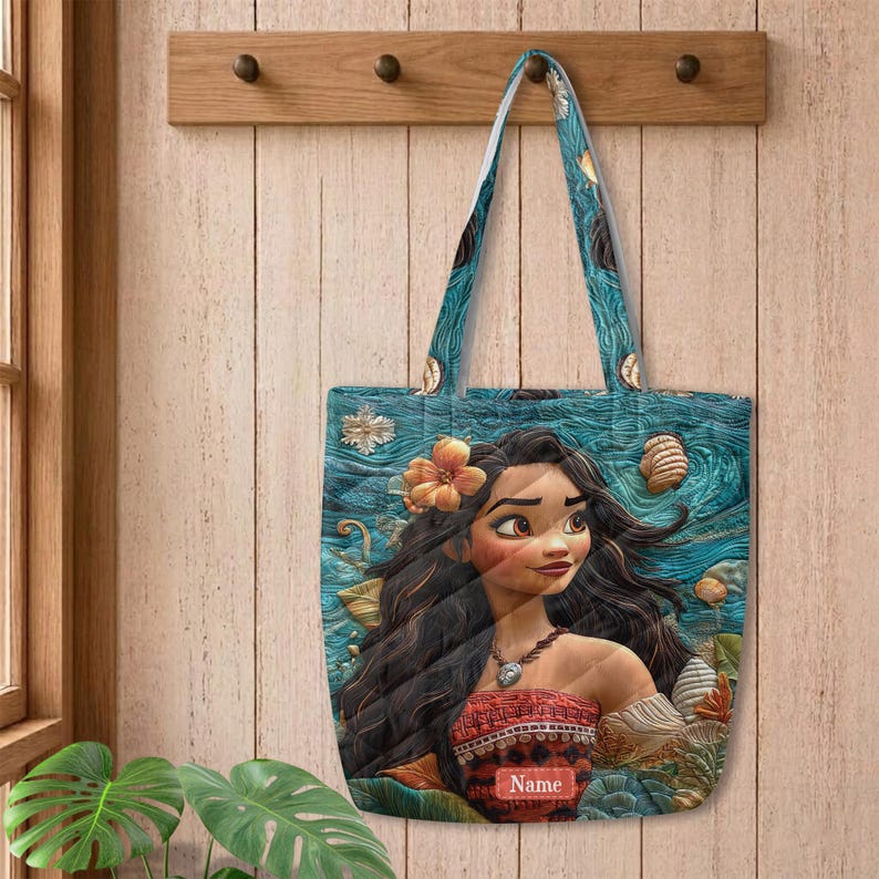 May include: A tote bag featuring a printed illustration of a character with long dark hair, a flower in their hair, and a necklace. The background shows a textured blue ocean pattern with seashells. The bag includes a rectangular label with the text Name.