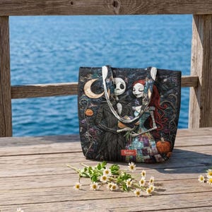 May include: A quilted tote bag featuring a printed illustration of two skeletal figures in a dark landscape with a crescent moon and a pumpkin. The bag has two shoulder straps and a small rectangular red label with the word Name printed on it.