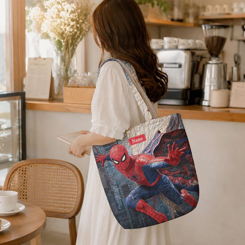 May include: A quilted tote bag featuring a graphic print of the superhero Spider-Man in a dynamic pose against a city background. The bag includes a rectangular red label with the word Name printed in white text on the upper portion of the design.