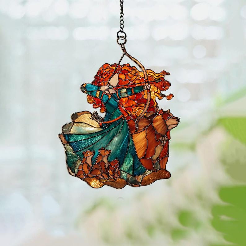 May include: Stained glass suncatcher featuring a woman with fiery red hair aiming a bow and arrow. She is wearing a teal and blue dress, with a bear and three cubs. The suncatcher is suspended by a chain.