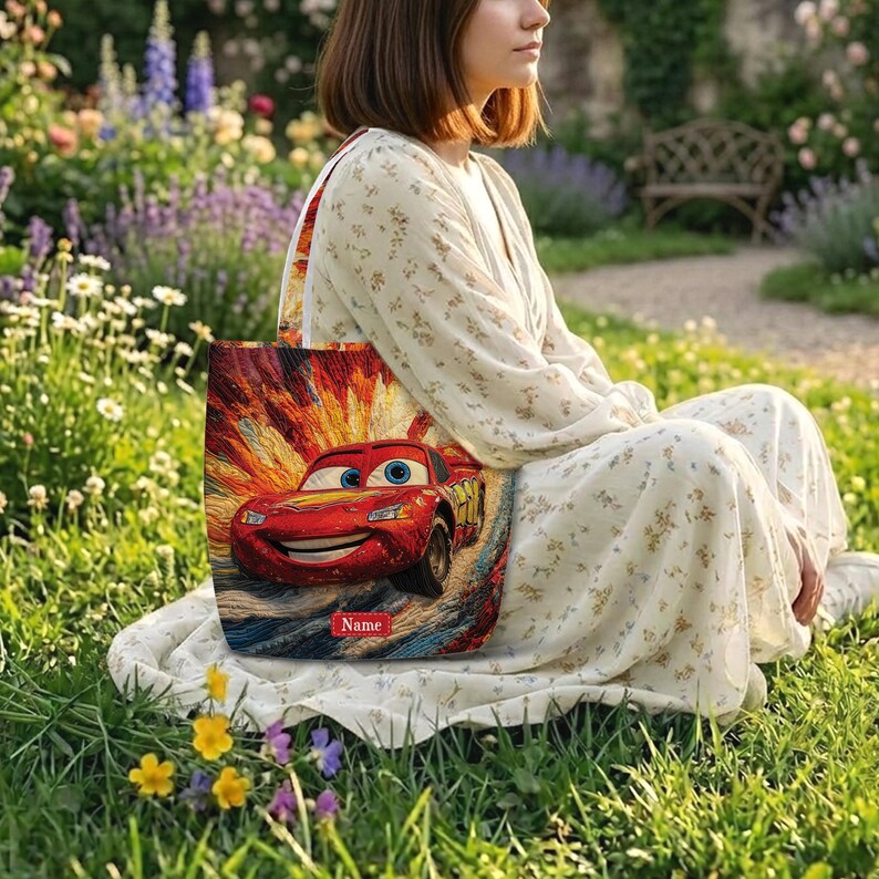 May include: A tote bag featuring a vibrant, textured illustration of a red race car with blue eyes and a wide smile. The background shows a dynamic orange and yellow burst effect. A small red rectangular label with the text Name is at the bottom.